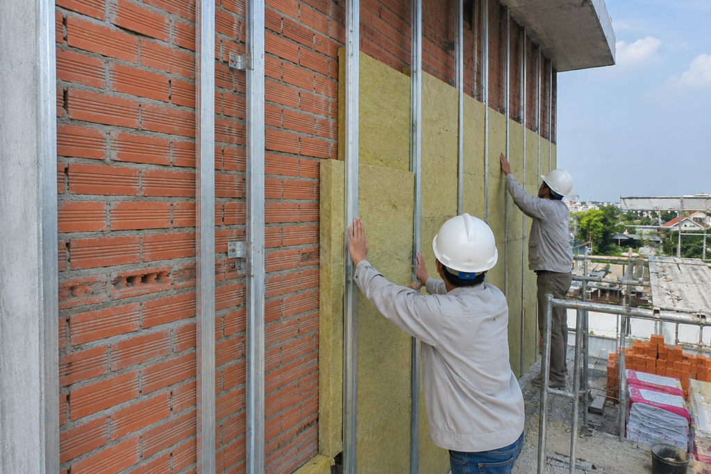 Đội ngũ thợ thi công lắp đặt tấm bông khoáng cách nhiệt chống nóng cho hệ tường gạch thô ngoài trời.